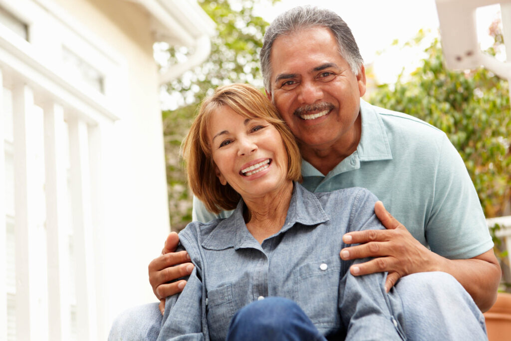 A close-up of a smiling senior couple. The man and woman are wearing light blue shirts and are seated. The woman is in front of the man, and the man's hands are embracing her shoulders. Out-of-focus bushes are visible in the background.